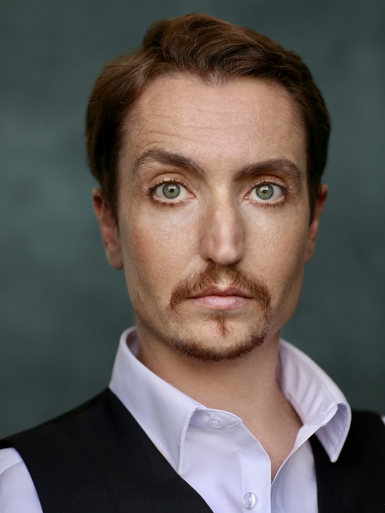 Head and shoulder photo of Cloud Quinn looking straight into the camera with a quizzical expression, his right eyebrow slightly raised. He has short brown hair and goatee and green-blue eyes. He wears a black waistcoat over a white button up shirt with collar and stands in front of a teal background in a photographer’s studio.