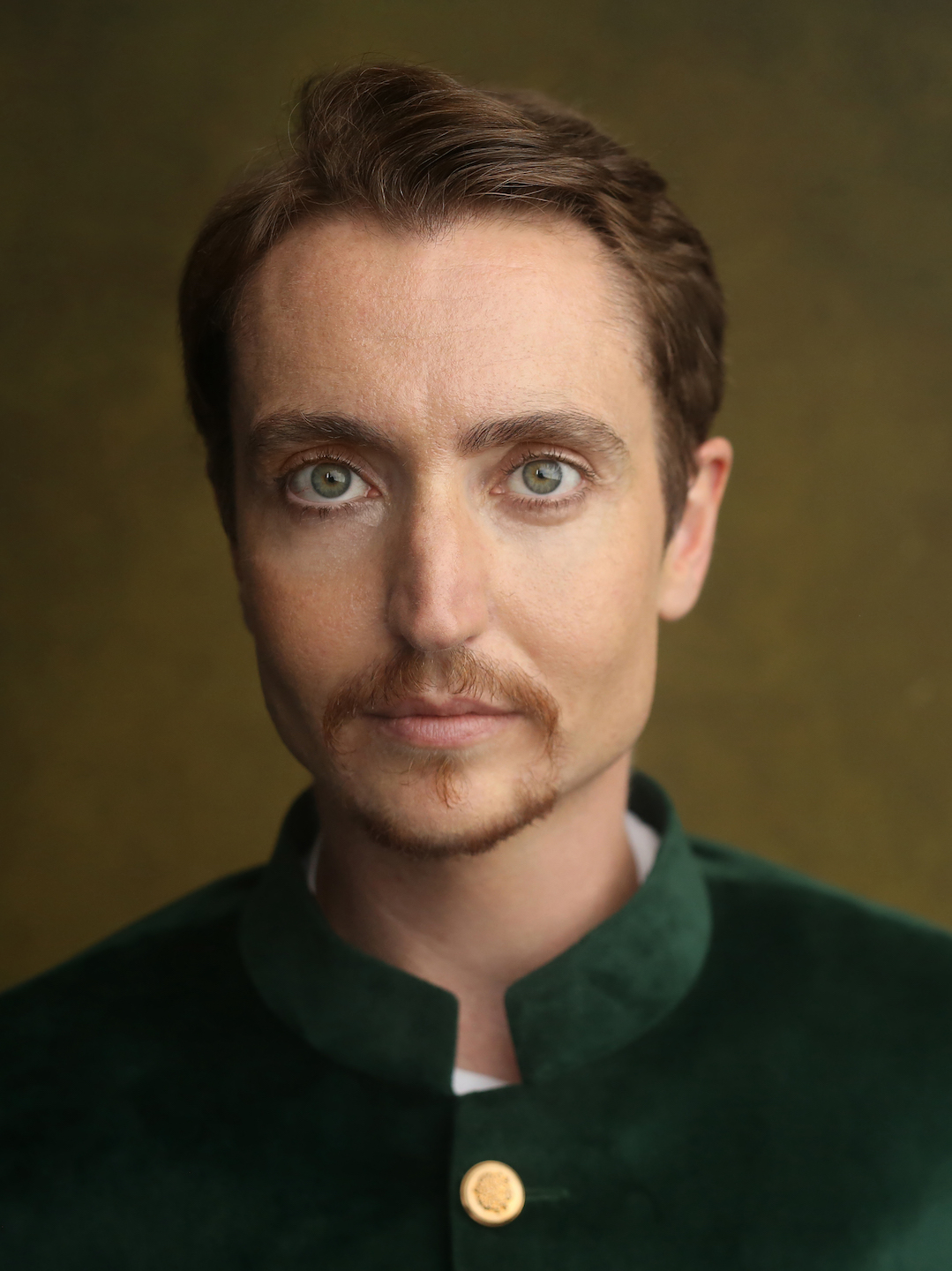 Head and shoulders photo of Cloud Quinn looking straight into the camera and smiling slightly. He has short brown hair and goatee and green-blue eyes. He wears a green velvet Nehru jacket with gold buttons and stands in front of a golden brown background in a photographer’s studio.